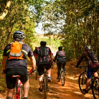 Imagem de 4 ciclistas de costas pedalando entre as árvores e o chão de terra do Parque Bike, no Granja Marileusa